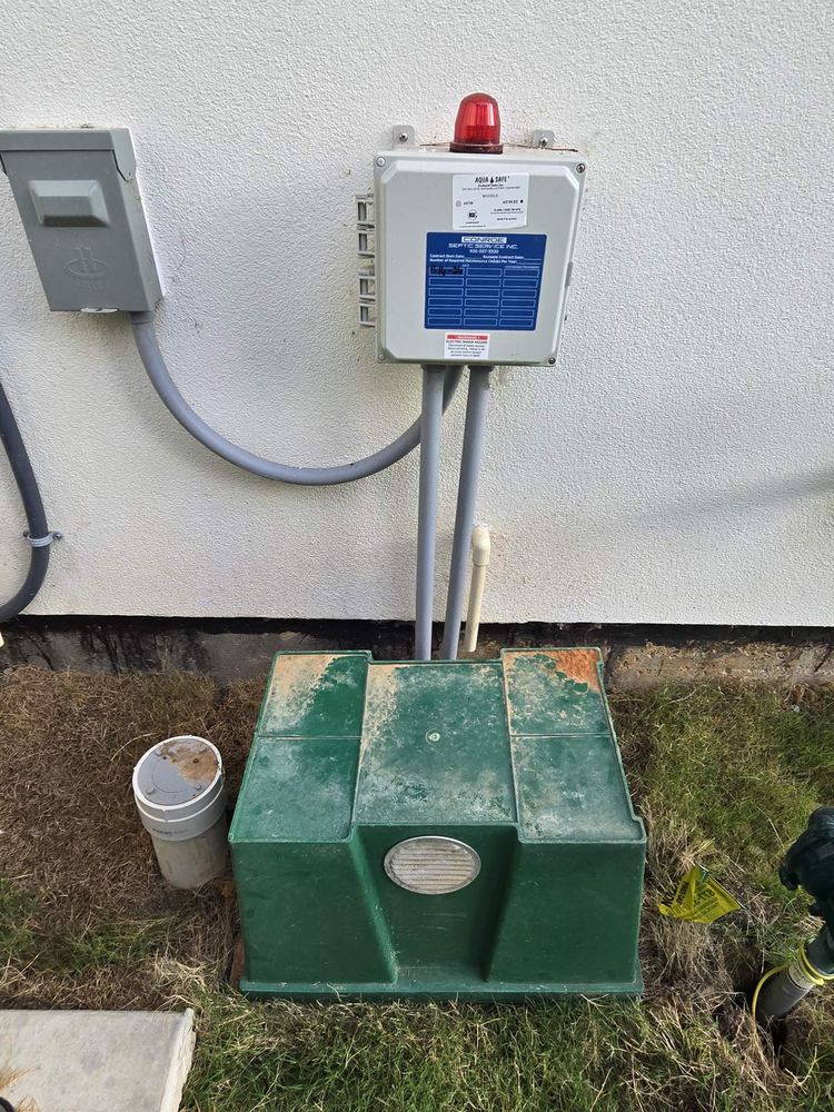 Green septic pump tank beside an exterior alarm panel, conduit, and a disconnect box