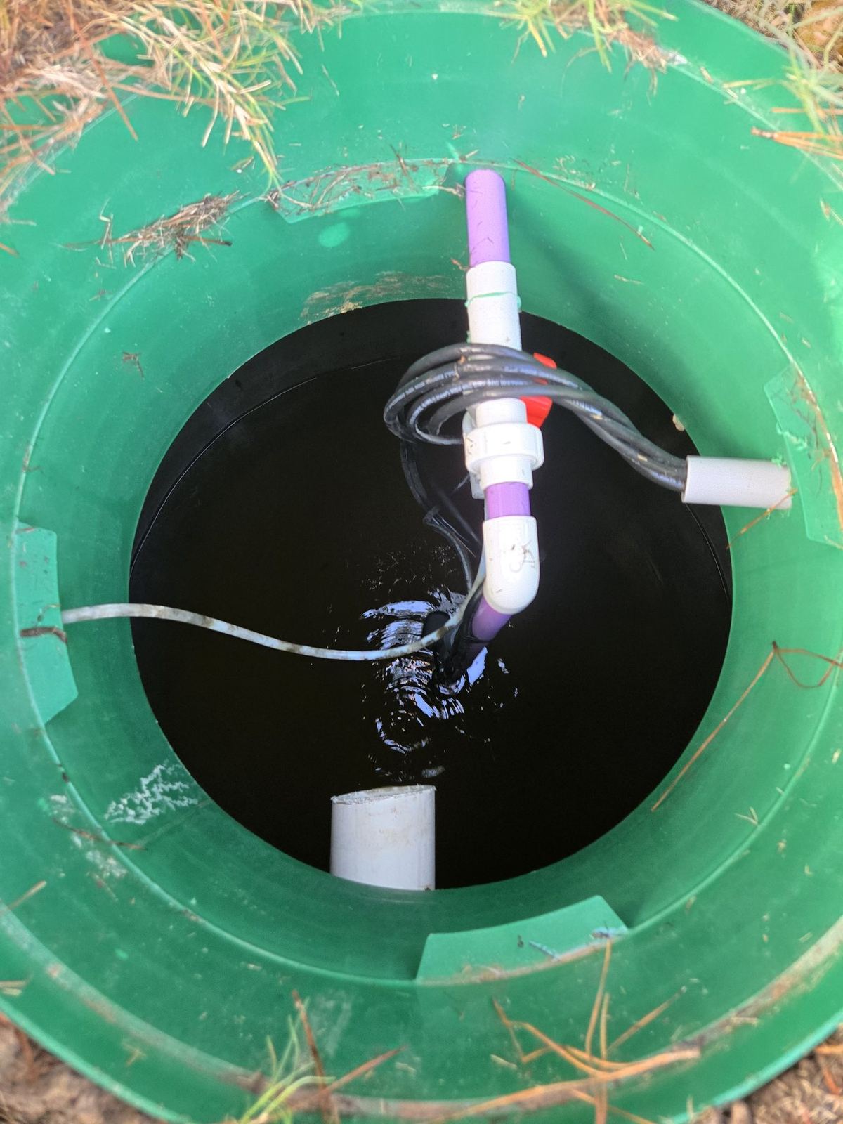 Open green septic tank riser with a pump, discharge pipe, and float-switch wires